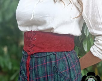 May include: A burgundy suede corset belt with a lace-up front detail. The belt is worn over a green and blue plaid skirt and a white peasant blouse. The belt is a wide style, designed to cinch the waist.