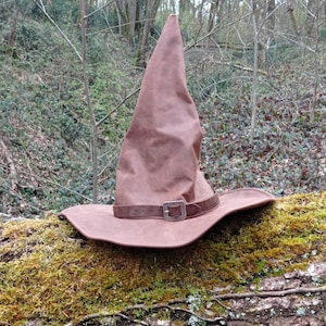 Op de afbeelding: Een bruine heksenhoed met een brede rand en een puntige kroon. De hoed heeft een bruine leren band met een zilverkleurige gesp. De hoed ligt op een met mos bedekte boomstam in een bos.