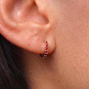 May include: A small gold hoop earring featuring a continuous row of tiny, vibrant red gemstones set along the outer edge. The circular earring is secured through the earlobe, showcasing a polished metallic finish and a series of faceted red stones.