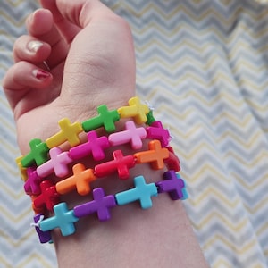 May include: A stack of colorful cross-shaped bracelets in shades of green, yellow, pink, red, orange, purple, and blue. The bracelets are strung together with white string and are worn on a wrist.