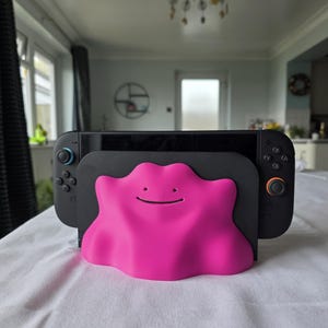 May include: A black Nintendo Switch console with attached controllers, held in a bright pink Ditto-shaped stand. The stand features a smiling face. The console and stand are on a white surface.