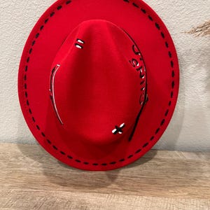 Peut inclure: Un chapeau fedora rouge vif avec un large bord. Le chapeau présente des coutures noires autour du bord et des coutures décoratives sur la couronne. Il comporte une bande argentée et le mot "SPRING" est écrit sur le côté.