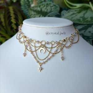 Può includere: Una collana girocollo a catene multiple color oro con delicate perline viola chiaro, un ciondolo centrale a mezzaluna e una piccola stella pendente sul fondo. La collana riporta la scritta @crystal.jwlr al centro del design.