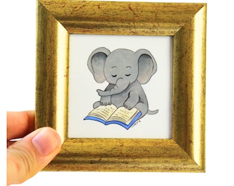 Peut inclure: Une petite œuvre d'art carrée, encadrée en or, représentant un éléphant de dessin animé gris lisant un livre bleu. L'éléphant a les yeux fermés. L'œuvre est tenue par une main.