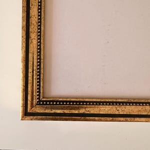 May include: A corner of a gold picture frame with a black accent. The frame has a textured, ornate design with small, evenly spaced dots along the inner edge. The frame is set against a white background.