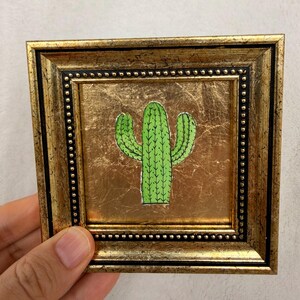 Peut inclure: Petite &oelig;uvre d'art encadr&eacute;e repr&eacute;sentant une illustration de cactus vert sur fond dor&eacute;. Le cadre est dor&eacute; avec une bordure noire perl&eacute;e. L'&oelig;uvre est tenue par une main.