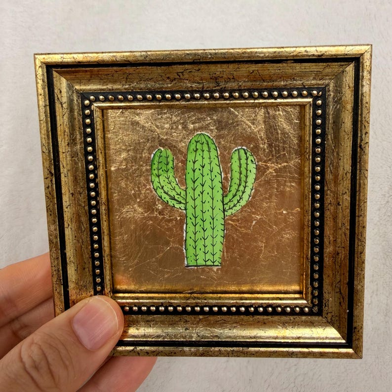 Peut inclure: Petite &oelig;uvre d'art encadr&eacute;e repr&eacute;sentant une illustration de cactus vert sur fond dor&eacute;. Le cadre est dor&eacute; avec une bordure noire perl&eacute;e. L'&oelig;uvre est tenue par une main.