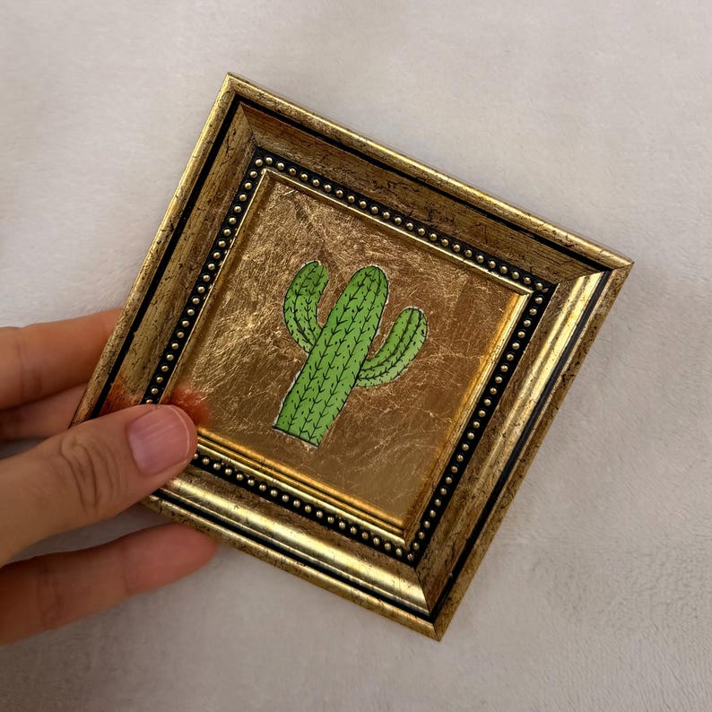 Peut inclure: Un cadre d&eacute;coratif carr&eacute; avec une finition dor&eacute;e m&eacute;tallique et une bordure int&eacute;rieure perl&eacute;e. Le centre pr&eacute;sente une illustration verte d'un cactus saguaro aux contours sombres, sur un fond dor&eacute; textur&eacute;.
