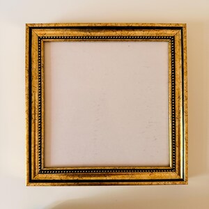 May include: Square gold picture frame with a detailed, ornate design. The frame has a textured, antique appearance with a black inner border and a white interior. The frame is empty, ready for a photo or artwork.