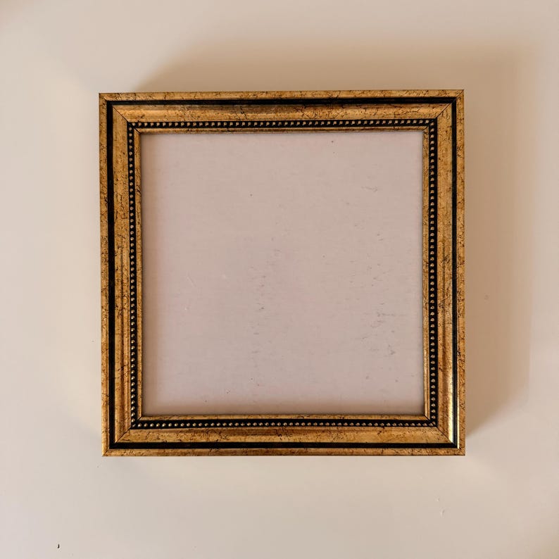 May include: Square gold picture frame with a black border and a decorative beaded edge. The frame has a vintage look and is empty, ready for a photo or artwork. The frame is displayed against a white background.
