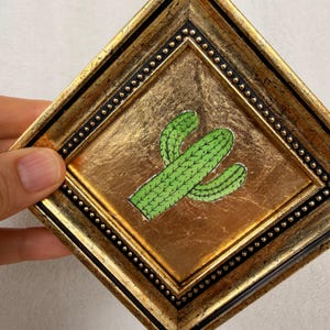 Peut inclure: Un cadre carr&eacute; dor&eacute; orn&eacute; avec une bordure int&eacute;rieure noire ponctu&eacute;e de petits clous dor&eacute;s. &Agrave; l'int&eacute;rieur, une illustration de cactus vert avec des &eacute;pines vert fonc&eacute; est centr&eacute;e sur un fond textur&eacute; en feuille d'or.