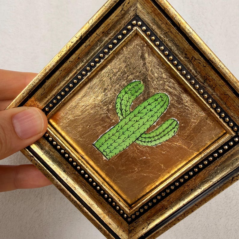 Peut inclure: Un cadre carr&eacute; dor&eacute; orn&eacute; avec une bordure int&eacute;rieure noire ponctu&eacute;e de petits clous dor&eacute;s. &Agrave; l'int&eacute;rieur, une illustration de cactus vert avec des &eacute;pines vert fonc&eacute; est centr&eacute;e sur un fond textur&eacute; en feuille d'or.