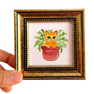 May include: A framed watercolor painting of an orange cat in a red flower pot surrounded by green leaves. The artwork is set in a gold-colored frame with a beaded border. The painting is held by a hand.