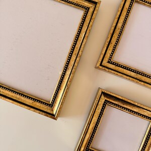 May include: Overhead shot of three gold picture frames with black accents. The frames have a rectangular shape with a white interior. The frames are arranged at different angles against a white background.