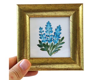 Könnte beinhalten: Ein kleiner, gerahmter Aquarellmalerei mit blauen Blumen und grünen Blättern auf weißem Hintergrund. Das Gemälde befindet sich in einem goldfarbenen Holzrahmen. Das Kunstwerk wird von einer Hand gehalten.