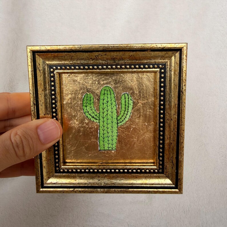 Peut inclure: Une &oelig;uvre d'art murale carr&eacute;e repr&eacute;sentant une illustration de cactus vert centr&eacute;e sur un fond en feuille d'or. L'&oelig;uvre est encadr&eacute;e dans un cadre dor&eacute; d&eacute;coratif avec une bordure int&eacute;rieure noire et un d&eacute;tail perl&eacute; sur le bord.