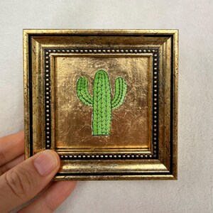 Peut inclure: Petite &oelig;uvre d'art encadr&eacute;e repr&eacute;sentant une illustration de cactus vert sur fond dor&eacute;. Le cadre est dor&eacute; avec une bordure perl&eacute;e. L'&oelig;uvre est tenue par une main.