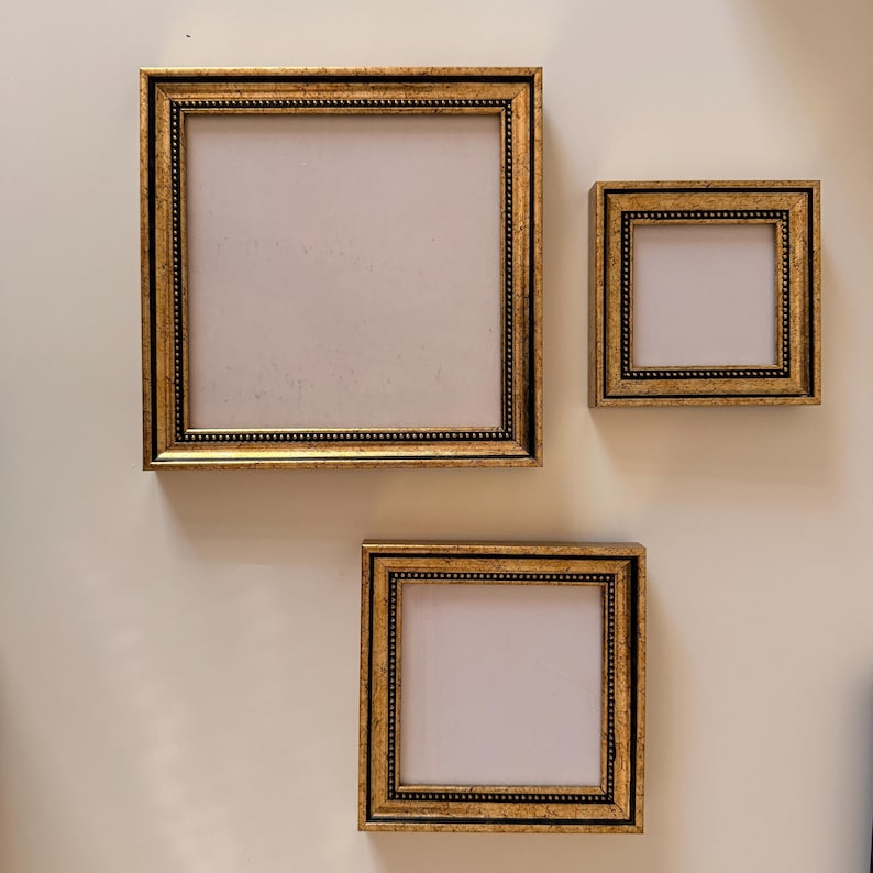 May include: Three gold-colored picture frames of varying sizes are mounted on a light-colored wall. The frames have a detailed, ornate design with a dark border and a beaded inner edge. The frames are empty, with a white interior.