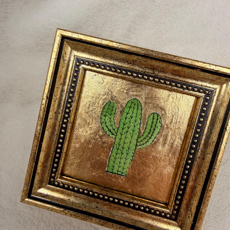 Peut inclure: Tableau carr&eacute; avec une illustration de cactus vert sur fond dor&eacute;. Le cadre est dor&eacute; avec une bordure int&eacute;rieure noire et de petits perles d&eacute;coratives. L'&oelig;uvre est une pi&egrave;ce d&eacute;corative.