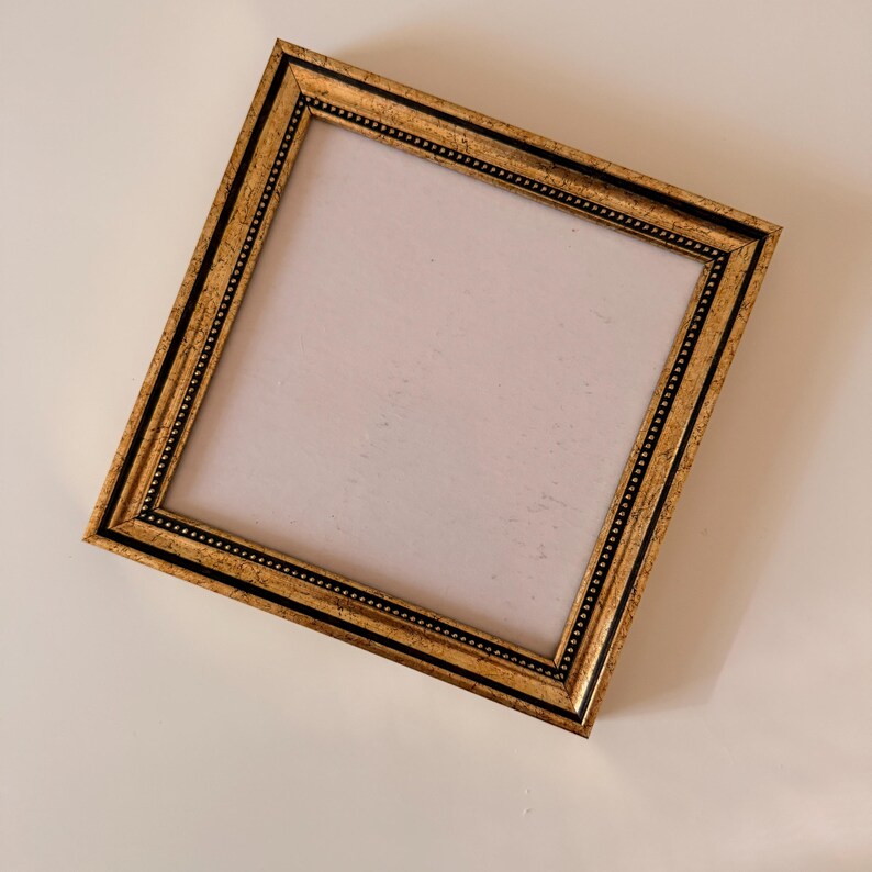 May include: A gold-colored picture frame with a black border and a white interior. The frame has a decorative beaded pattern along the inner edge. The frame is square and angled, showcasing its ornate design.