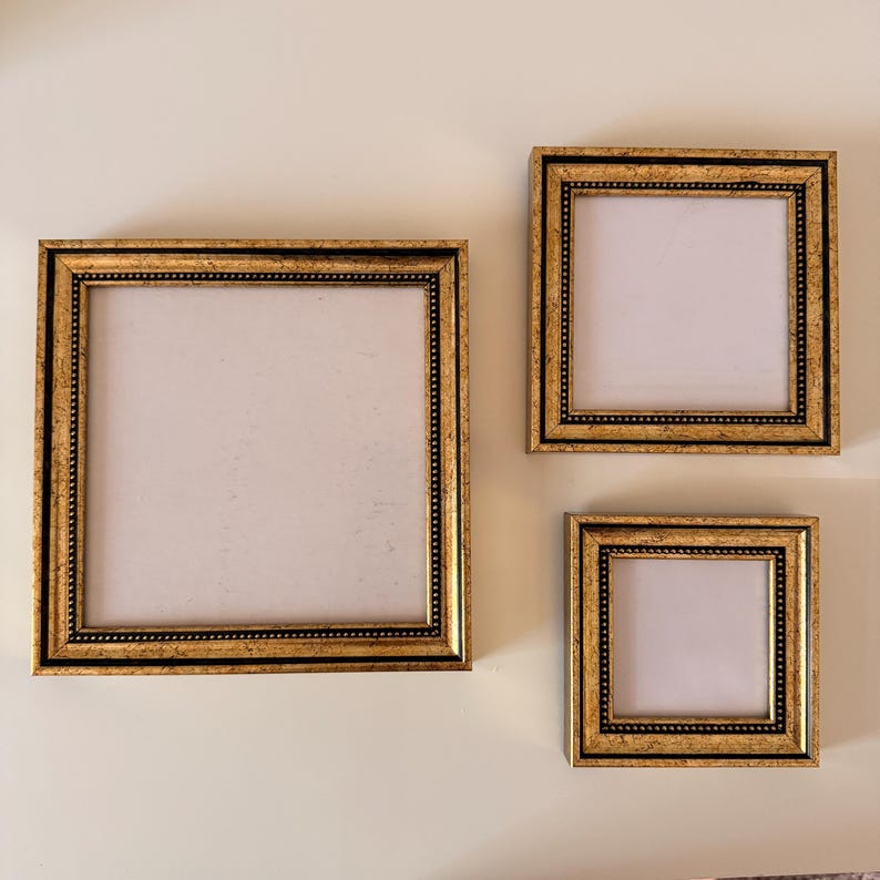 May include: Three gold-colored picture frames of varying sizes are displayed against a neutral-toned wall. The frames have a square shape with ornate detailing and a black border. The frames are empty, ready for photos or artwork.