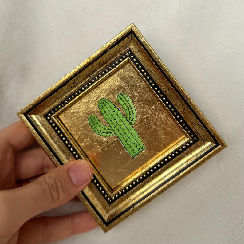Peut inclure: Petite &oelig;uvre d'art carr&eacute;e encadr&eacute;e, repr&eacute;sentant un cactus vert sur fond dor&eacute;. Le cadre est dor&eacute; avec un bord int&eacute;rieur noir et un bord perl&eacute;. L'&oelig;uvre est tenue par une main.