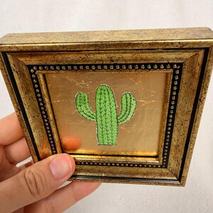 Peut inclure: Petite &oelig;uvre d'art carr&eacute;e encadr&eacute;e en or, repr&eacute;sentant un cactus vert dessin&eacute; &agrave; la main sur un fond dor&eacute;. Le cadre a un aspect textur&eacute; et vieilli, avec un bord int&eacute;rieur noir et des perles d&eacute;coratives.