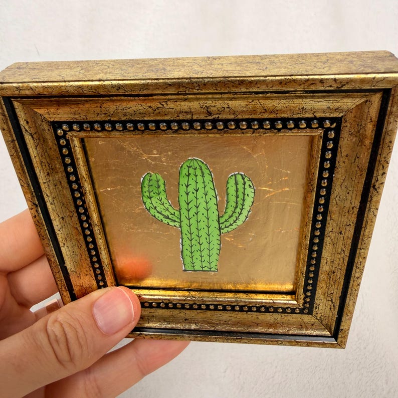 Peut inclure: Petite &oelig;uvre d'art carr&eacute;e encadr&eacute;e en or, repr&eacute;sentant un cactus vert dessin&eacute; &agrave; la main sur un fond dor&eacute;. Le cadre a un aspect textur&eacute; et vieilli, avec un bord int&eacute;rieur noir et des perles d&eacute;coratives.