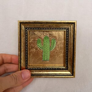 Peut inclure: Petite &oelig;uvre d'art encadr&eacute;e carr&eacute;e avec une illustration de cactus vert. Le cadre est dor&eacute; avec une finition textur&eacute;e et une bordure int&eacute;rieure perl&eacute;e. Le cactus est centr&eacute; sur un fond dor&eacute;.