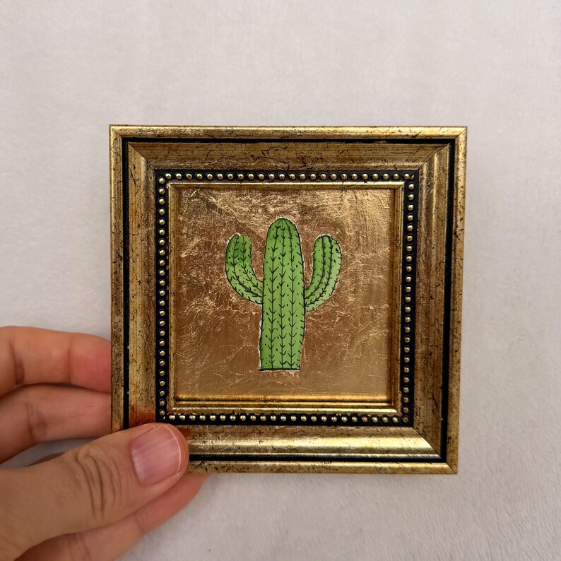 Peut inclure: Petite &oelig;uvre d'art encadr&eacute;e carr&eacute;e avec une illustration de cactus vert. Le cadre est dor&eacute; avec une finition textur&eacute;e et une bordure int&eacute;rieure perl&eacute;e. Le cactus est centr&eacute; sur un fond dor&eacute;.