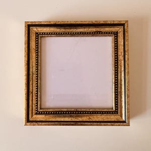 May include: Square gold picture frame with a black inner border and a beaded edge. The frame has a distressed finish, giving it a vintage look. The frame is empty, ready to display a photo or artwork.