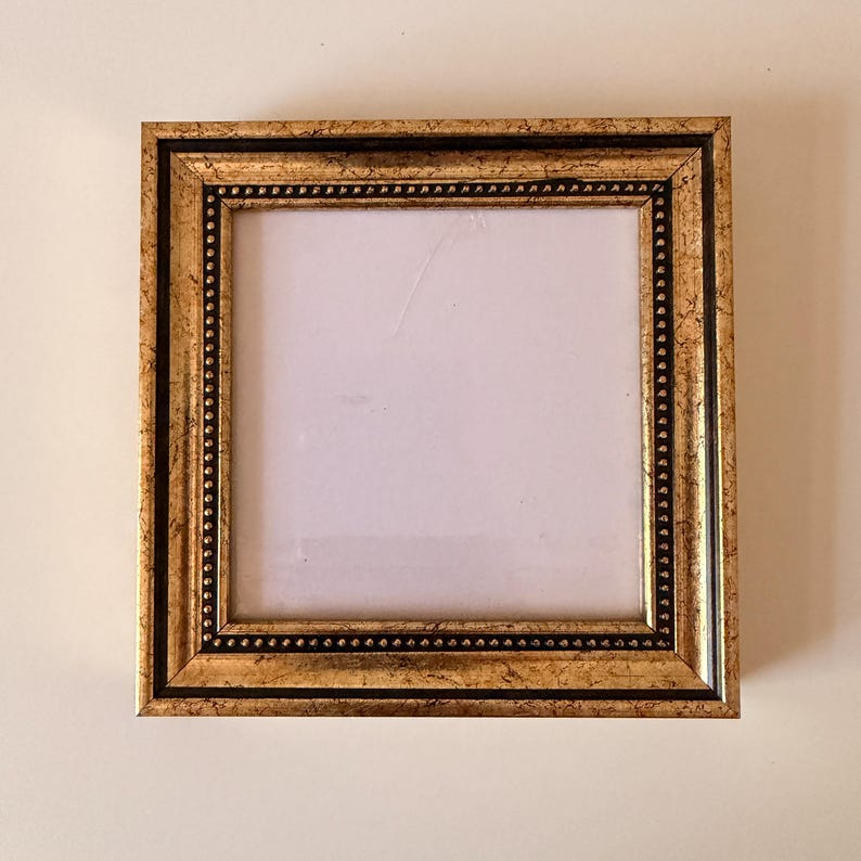 May include: Square gold picture frame with a black inner border and a beaded edge. The frame has a distressed finish, giving it a vintage look. The frame is empty, ready to display a photo or artwork.