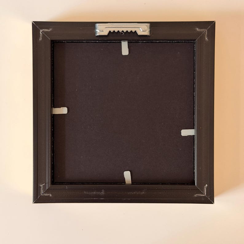 May include: The back of a square, black picture frame. The frame has a dark brown backing and white clips to hold the picture in place. A metal sawtooth hanger is attached to the top center of the frame for hanging.