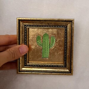 Peut inclure: Petite &oelig;uvre d'art carr&eacute;e encadr&eacute;e, repr&eacute;sentant un cactus vert sur fond dor&eacute;. Le cadre est de couleur or fonc&eacute; avec un bord int&eacute;rieur perl&eacute;. L'&oelig;uvre est tenue par une main.