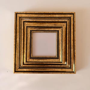 May include: A square, ornate picture frame with a gold finish and black accents. The frame has multiple layers, each with a beaded border. The center opening is white, ready for a photo or artwork.