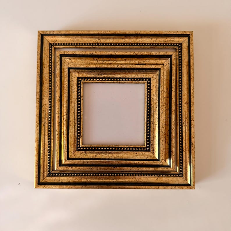 May include: A square, ornate picture frame with a gold finish and black accents. The frame has multiple layers, each with a beaded border. The center opening is white, ready for a photo or artwork.