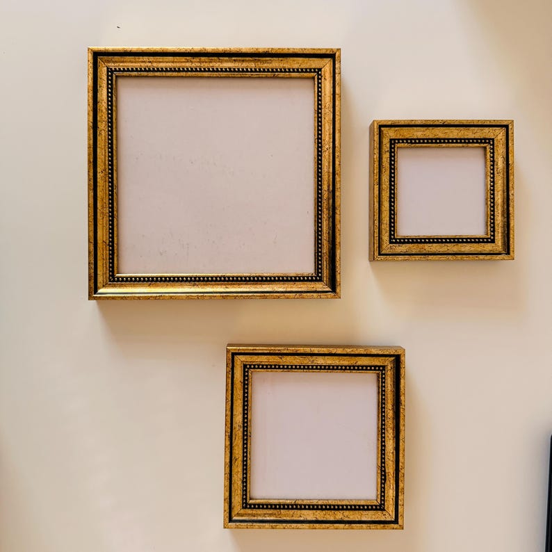 May include: Three gold-framed picture frames of varying sizes are displayed on a cream-colored wall. The frames have a detailed, ornate design with a black inner border. The frames are empty, with a blank, off-white interior.
