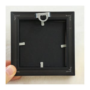 Peut inclure: Un cadre photo carr&eacute; noir avec un dos noir. Le cadre a un crochet en m&eacute;tal en haut et des clips en m&eacute;tal pour maintenir le dos en place. Le cadre est tenu par une personne.