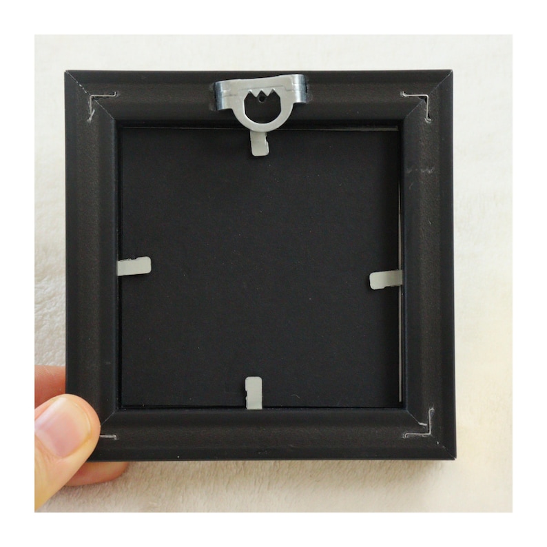 Peut inclure: Un cadre photo carr&eacute; noir avec un dos noir. Le cadre a un crochet en m&eacute;tal en haut et des clips en m&eacute;tal pour maintenir le dos en place. Le cadre est tenu par une personne.