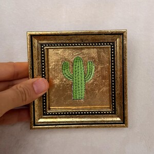 Peut inclure: Petite &oelig;uvre d'art carr&eacute;e encadr&eacute;e, repr&eacute;sentant un cactus vert sur fond dor&eacute;. Le cadre dor&eacute; est orn&eacute; d'un liser&eacute; int&eacute;rieur perl&eacute;. L'&oelig;uvre est tenue par une main.