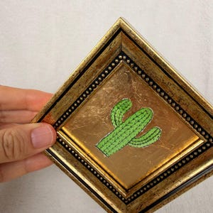 Peut inclure: Une &oelig;uvre d'art encadr&eacute;e repr&eacute;sentant un cactus vert sur fond dor&eacute;. Le cadre dor&eacute; est bord&eacute; de perles noires. L'&oelig;uvre est tenue par une main.