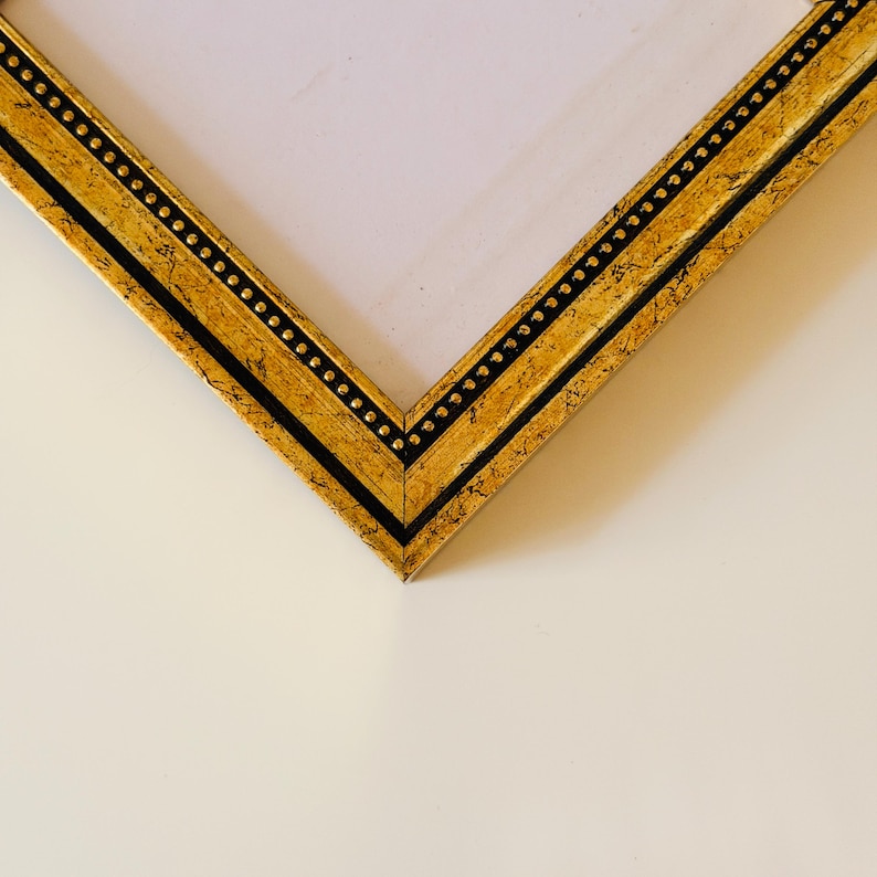 May include: Close-up of a gold picture frame corner. The frame has a textured gold finish with black accents and a row of small, evenly spaced dots. The frame is angled, revealing a white interior.