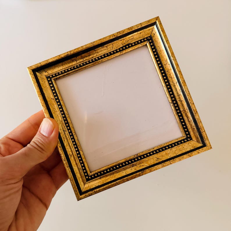 May include: Square gold picture frame with a black border and a beaded design. The frame is held at an angle, showcasing its ornate details and the blank space for a photo or artwork.
