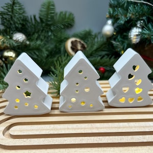 Könnte beinhalten: Drei weiße Keramik-Weihnachtsbaum-Kerzenhalter mit ausgeschnittenen Designs. Die Kerzenhalter stehen auf einem Holztablett, und der Hintergrund zeigt einen Weihnachtsbaum mit Ornamenten. Die Ausschnitte umfassen Sterne, Kreise und Herzen.