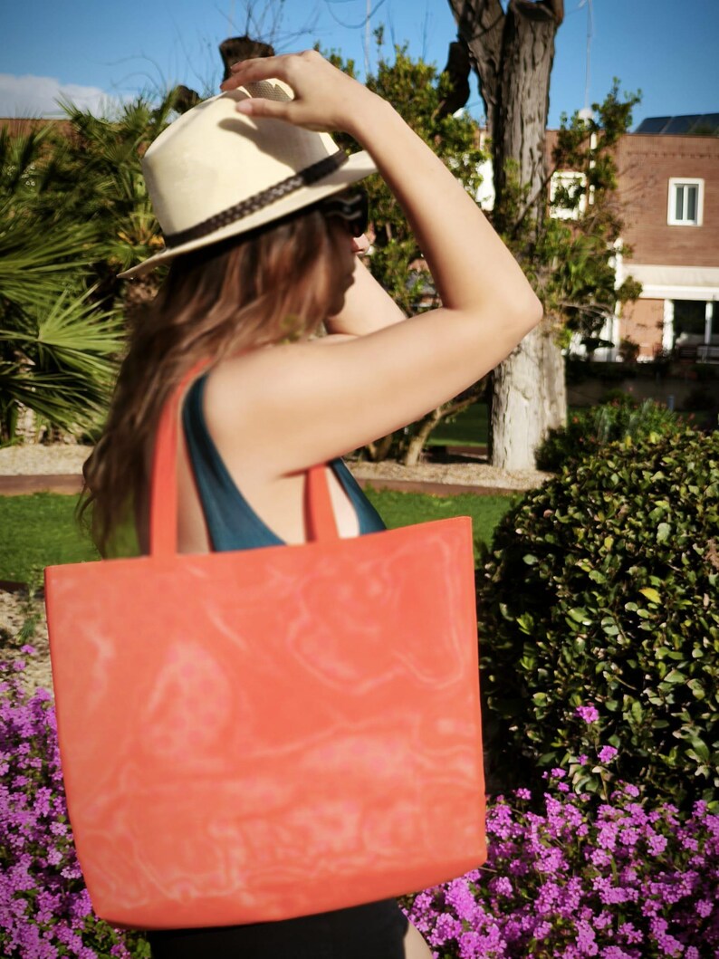May include: A woman wearing a straw hat and sunglasses is carrying a large orange tote bag. The bag has a faded design on it.