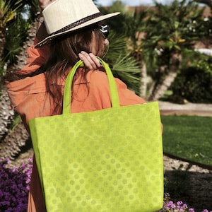 May include: A woman wearing a straw hat and sunglasses carries a green tote bag with white polka dots. The bag is made of a textured fabric and has a wide, flat base.