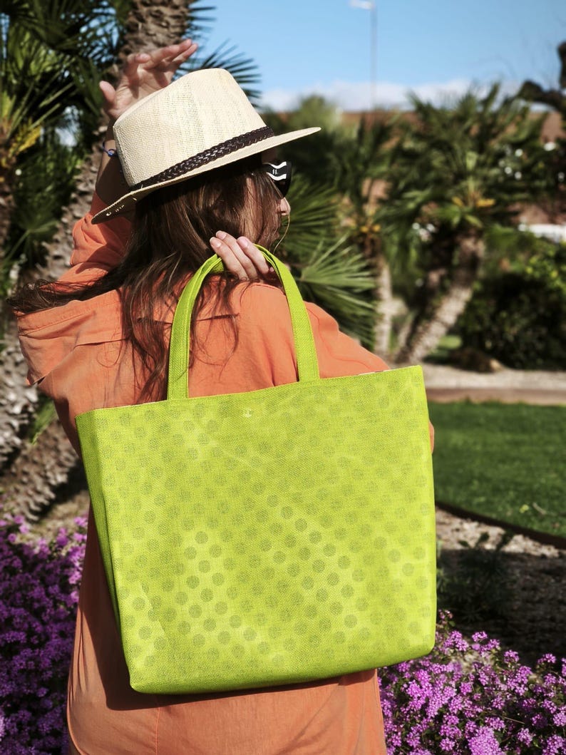May include: A woman wearing a straw hat and sunglasses carries a green tote bag with white polka dots. The bag is made of a textured fabric and has a wide, flat base.