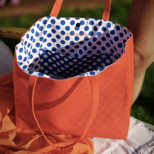 May include: Orange canvas tote bag with blue and white polka dot lining. The bag has a small pocket on the inside and the text "by Conchita" is printed on the lining.