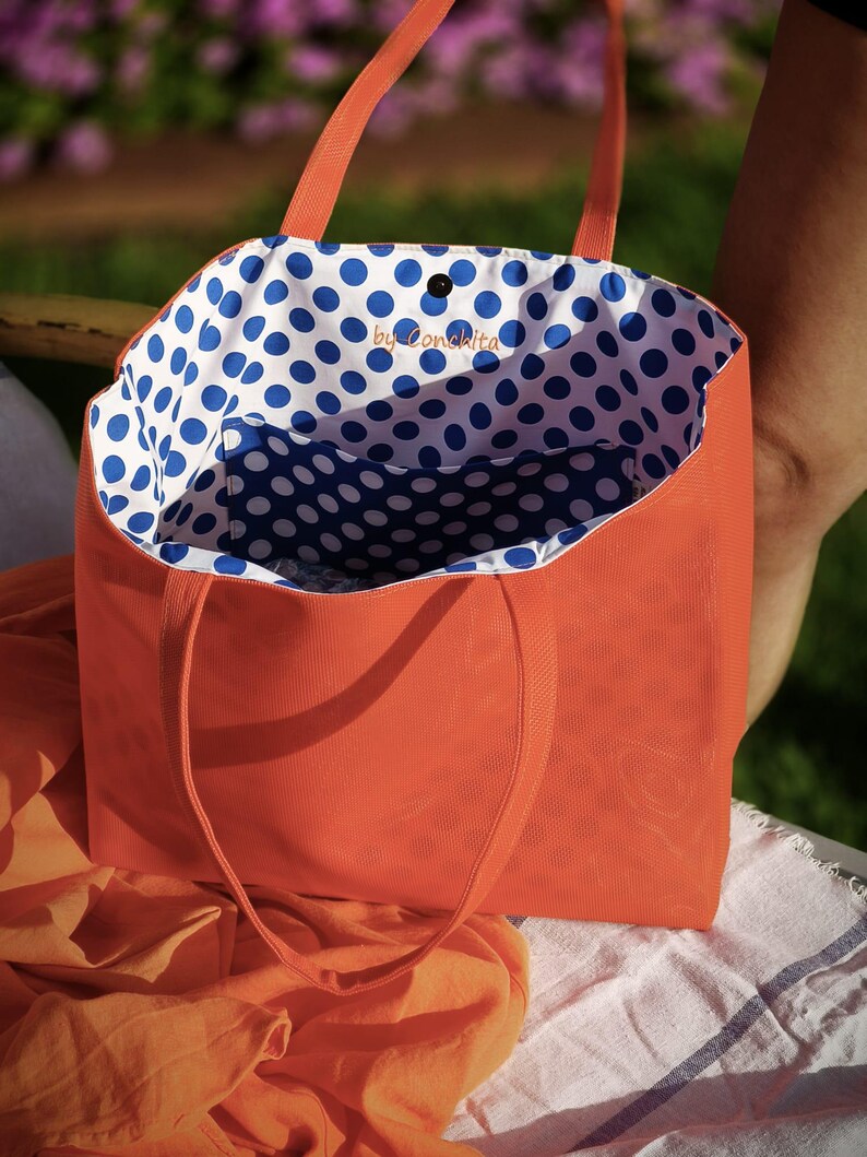 May include: Orange canvas tote bag with blue and white polka dot lining. The bag has a small pocket on the inside and the text "by Conchita" is printed on the lining.