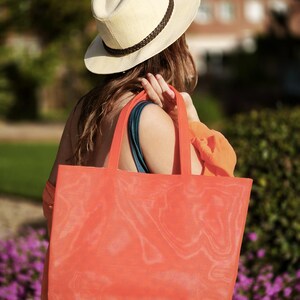 May include: A woman wearing a straw hat and a bright orange mesh tote bag. The bag is large and has a wide opening. The woman is walking away from the camera.